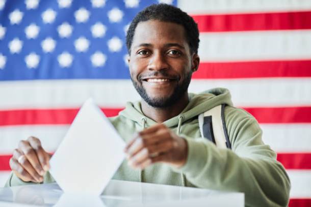 Portrait of smiling black man putting ballot in bin against American flag